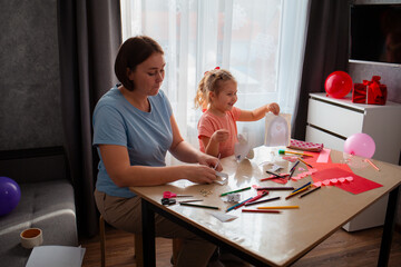 Wide shot of kindergarten teacher plays and teaches cute little happy girl how to make paper cards for Mother's Day and Valentine's Day. Woman and child sitting at the table