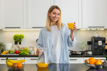 Middle-aged woman pours orange juice into glass and drinks for breakfast in home kitchen for daily...