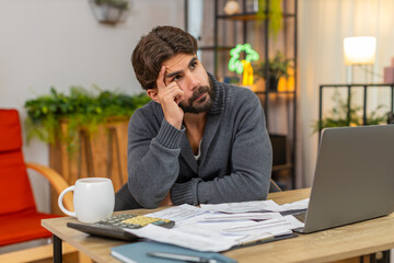 Indian man at home leans on hands holds head sees unpaid bills on table by calculator and laptop....