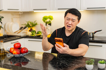 Asian man in home kitchen looks at orange smartphone with shocked expression after online game loss...