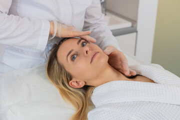 Young woman lies in clinic chair while cosmetologist in gloves examines facial skin before...