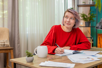 Young woman carefully writing notes from financial documents sitting at home table, serious and concentrated. Caucasian girl demonstrating focused, attentive business working process and productivity.