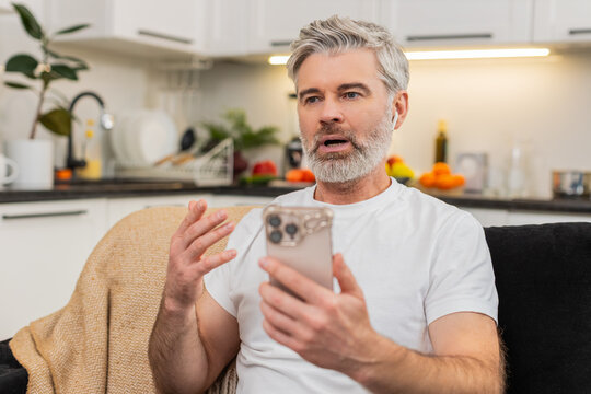 Middle-aged man at home learns phrases on smartphone app with earphones repeats words and hears feedback. Mature guy on sofa pauses and smiles sees high score feels big progress and keeps study flow.
