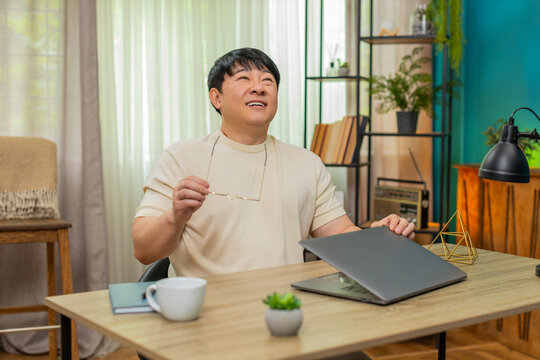 Asian man sitting at home table finishes work, closes laptop, removes glasses, rests back in chair with happy sigh. Chinese guy feels relief, savors completed goals and earned break after long hours.