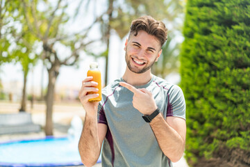 Fototapeta premium Young handsome sport man holding an orange juice at outdoors and pointing it