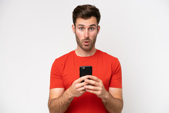 Young caucasian man isolated on white background looking at the camera while using the mobile with surprised expression