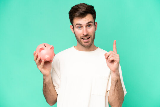 Young caucasian man holding piggybank isolated on green background pointing up a great idea