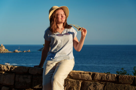Portrait of a young woman squinting at the bright sunset sun, holding her sunglasses and wearing a straw hat by the seaside during holidays.