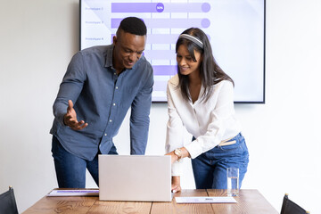Diverse coworkers leaning over table in boardroom analyzing data on laptop, screen with charts