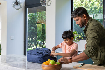 Indian father and son standing at marble island countertop unpacking blue backpack, copy space © wavebreak3