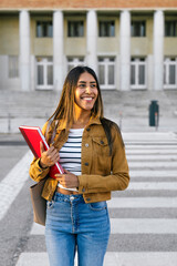 Young woman confidently walking on campus, holding red notebooks, representing higher education...