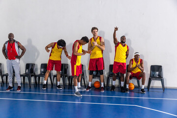 Diverse male basketball team standing on basketball court holding balls, coach blowing whistle
