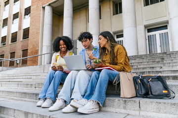 Multiracial university students using a laptop, chatting, and eating on campus steps while taking a...