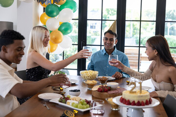 Diverse friends wearing party hats around wooden table celebrating birthday with cake, balloons