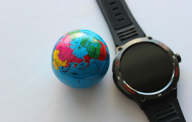 A model of a globe next to a round smartwatch on a white background with a blank space for text on the side. Photo of watch and globe illustrating the topic of time and distance
