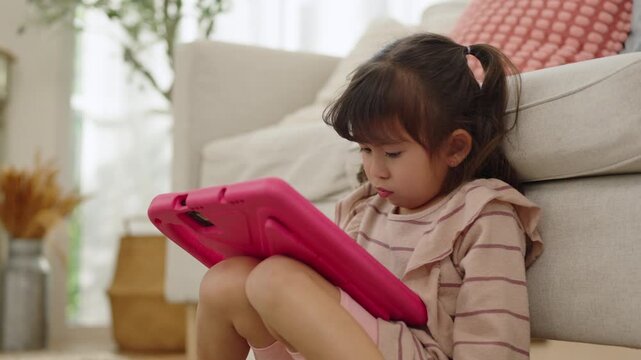 Asian young girl sitting at home and focusing on playing a game on a tablet, a female child enjoying calm and quiet digital screen time.