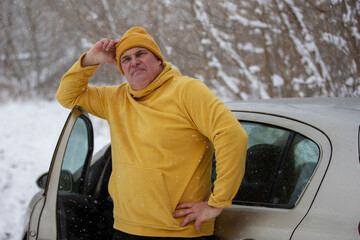 Man in yellow hoodie and beanie standing beside car in snowy landscape during winter