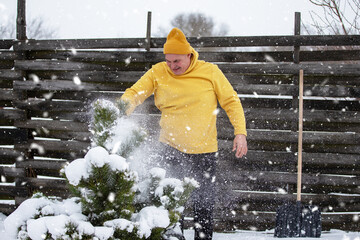 Man in yellow sweater shoveling snow from a tree in a snowy garden during winter