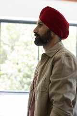 Obraz premium Indian man wearing red turban and jacket by window in natural light, gazing at foliage, copy space