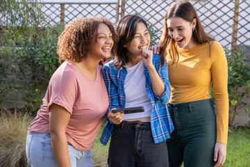 Diverse female friends laughing while looking at black handheld device in backyard by lattice fence © wavebreak3