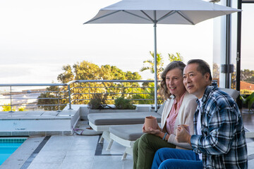 Diverse senior couple relaxing at poolside holding mugs near umbrella with ocean view, copy space