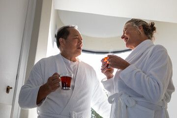 Diverse couple standing near curved-top window in spa suite holding steaming tea mugs