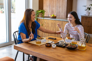 Mother and daughter sitting at table in kitchen sharing salad with chopsticks and chatting. © wavebreak3