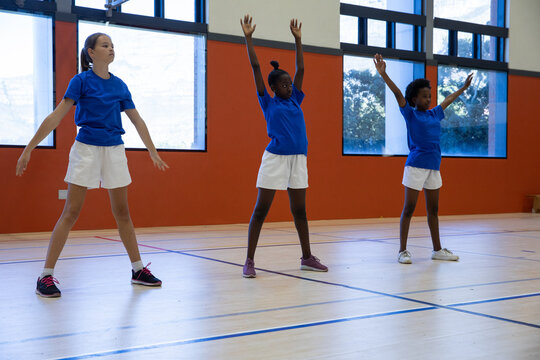 Diverse female students warming up in school gym on wooden court with lines and windows