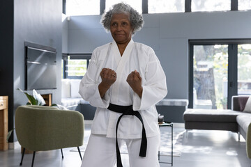 Naklejka premium Senior african american woman holding fighting stance in living room wearing karate gi and belt