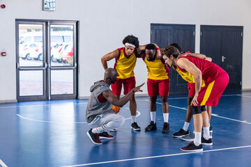 Diverse male basketball coach and team huddling on court holding orange ball directing play