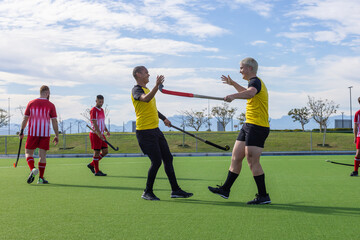 Male players in yellow jerseys celebrating victory on turf field with hockey sticks