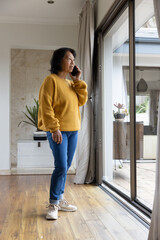 Senior Asian woman holding smartphone looking outside by sliding glass doors in living room