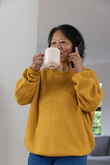 Senior asian woman in mustard sweater using smartphone, sipping from ceramic mug in living room