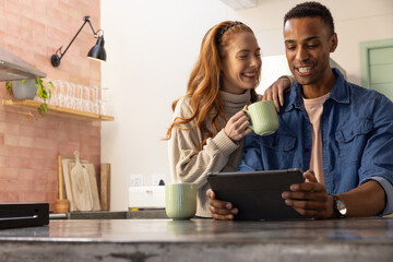 Diverse couple standing at kitchen island browsing tablet screen with green ceramic mugs