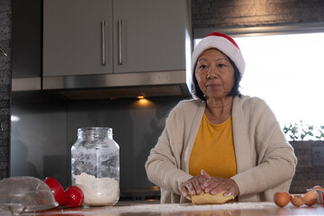 Senior Asian woman kneading dough on wooden countertop in kitchen with flour jar and Santa hat