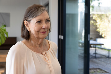 Senior woman standing in dining room with wooden table near sliding glass door, copy space