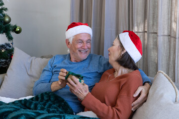 Senior couple exchanging gift box on sofa at home while wearing Santa hats near Christmas tree