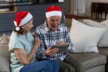 Senior couple sitting on sofa near kitchen cabinets pointing at tablet wearing Santa hats