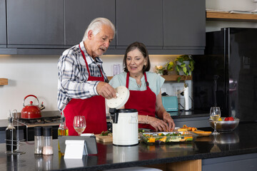 Senior couple wearing red aprons operating food processor and slicing vegetables at kitchen island