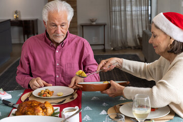 Senior couple spooning roast bird and vegetables onto plate on festive tablecloth in dining room