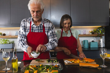 Senior couple cooking at kitchen island wearing red aprons, sprinkling herbs and slicing carrots