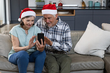 Senior couple wearing Santa hats using smartphone on grey leather couch in open kitchen at home