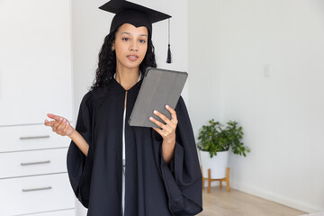Female teenager holding tablet, gesturing at home with graduation gown mortarboard cap and plant