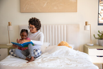 African american mother with daughter reading kids book on tufted bed with teddy bear, copy space