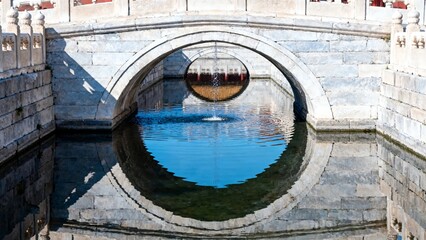 Stone arch bridge over calm water with circular reflection in a tranquil garden setting