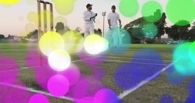 Cricket batsmen walking onto crease after call, preparing to bat beside stumps under colorful bokeh