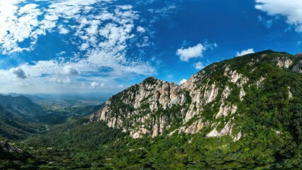 Naklejka premium A panoramic view of a rugged mountain range with lush greenery under a bright blue sky with scattered clouds