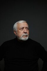 Older man with white beard and gray hair looks thoughtfully while wearing black clothing in a dark room