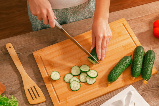 Preparing a salad. A woman's hands use a knife to slice cucumbers into thin slices on a wooden board. Top view.