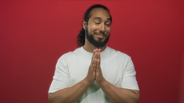 Smiling man in white t shirt with hands pressed together in prayer gesture in red studio; playful plea.